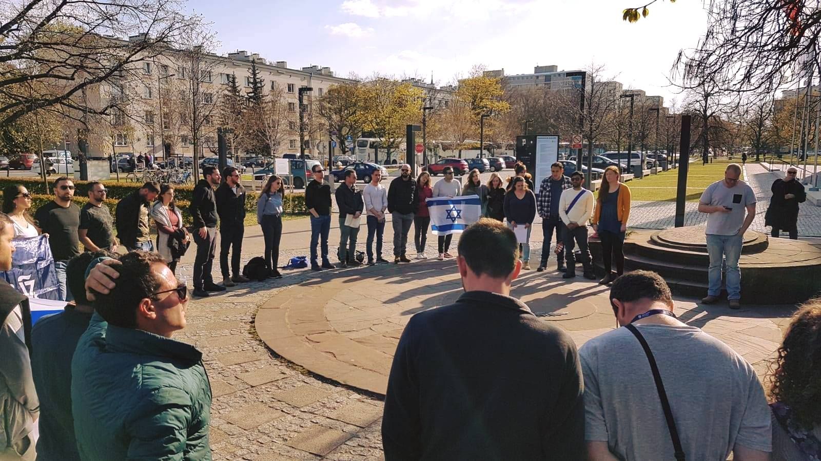 מעגל סטודנטים עומד בכיכר עירונית בוורשה עם דגל ישראל בשמש של בוקר