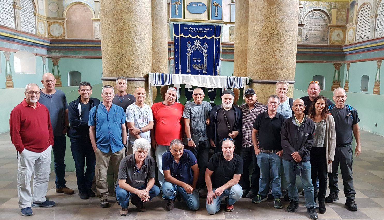 משלחת גברים בוגרים מצטלמת בתוך בית כנסת היסטורי לצד עמודי עץ מעוטרים