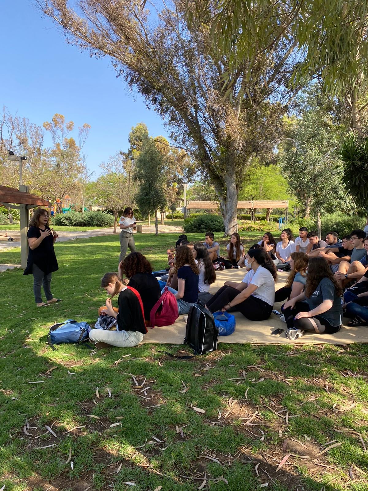 מדריכה עומדת מול קבוצת תלמידים היושבים על שמיכה בדשא הגן