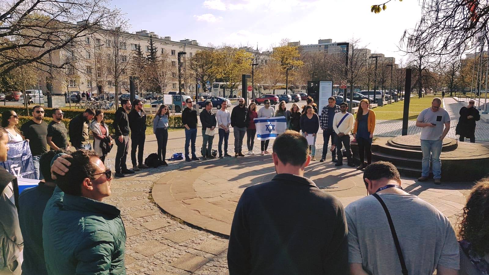 מעגל סטודנטים עומד בכיכר עירונית בוורשה עם דגל ישראל בשמש של בוקר