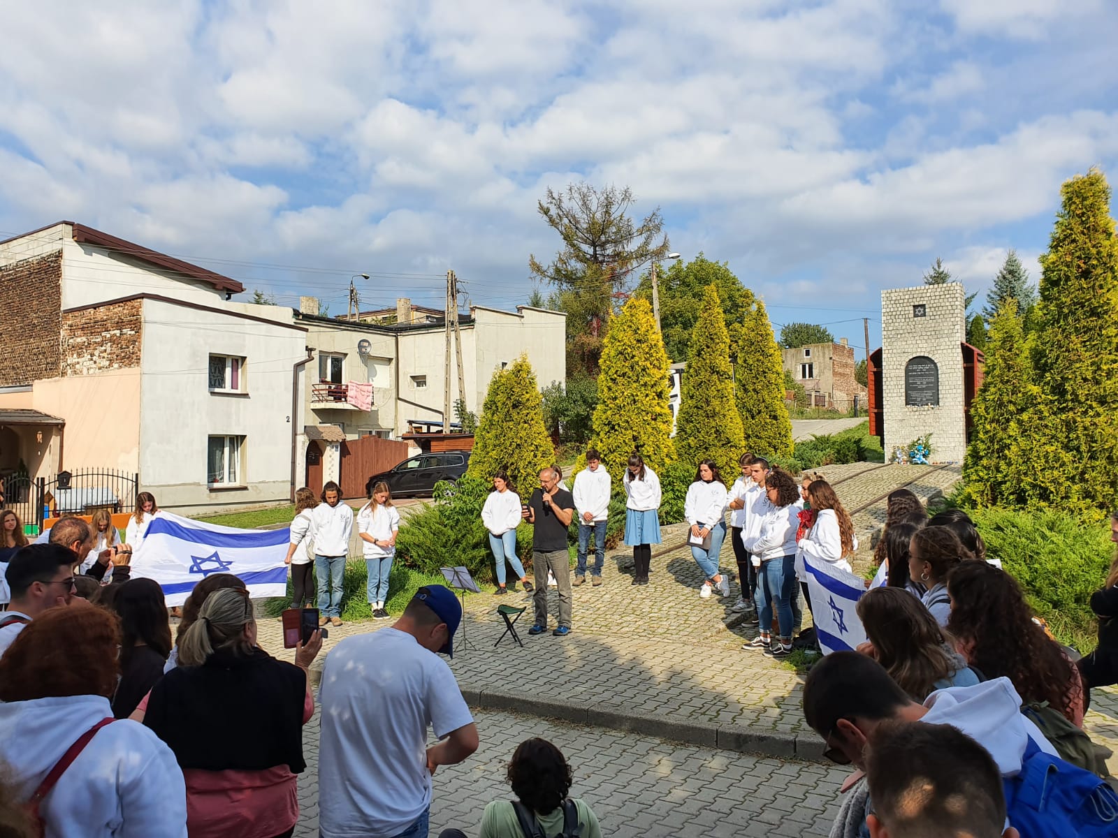 בני נוער בחולצות לבנות מקיימים טקס זיכרון עם דגל ישראל ליד אנדרטה בעיירה פולנית