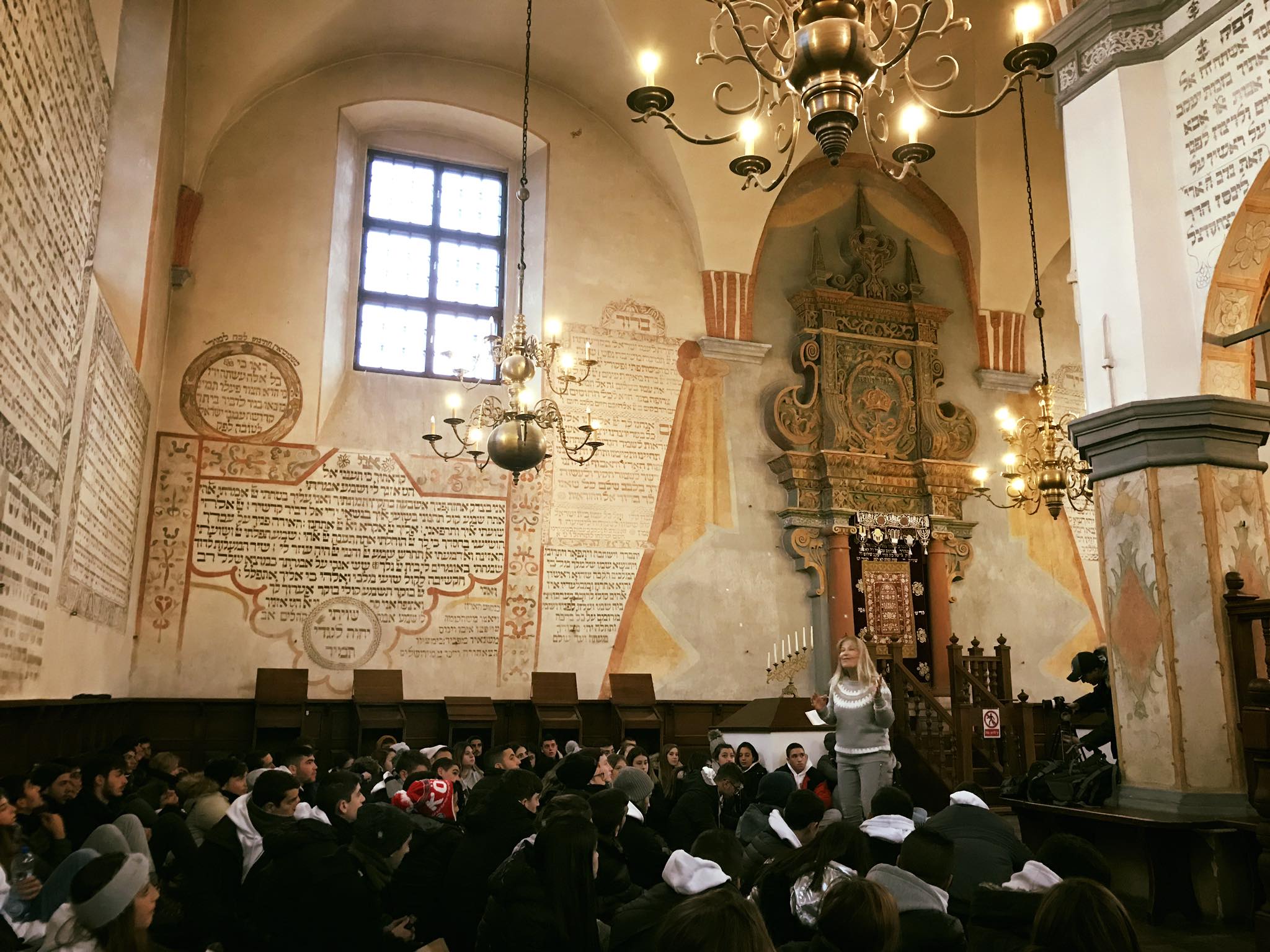 קבוצת תלמידים מקשיבה למדריכה בתוך בית כנסת היסטורי מקושט ציורי קיר